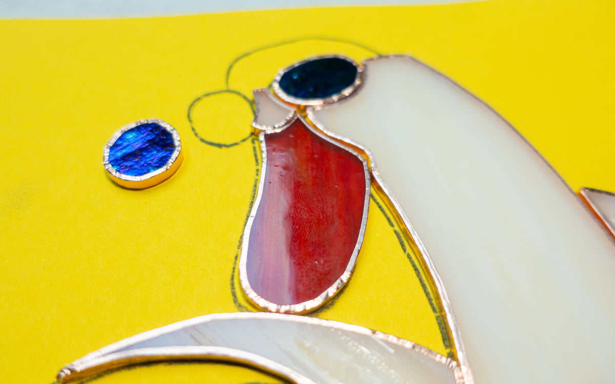 Close-up detail of banana duck stained glass suncatcher face — blue glass sunglasses, red beak, copper foil soldering, studio process photo