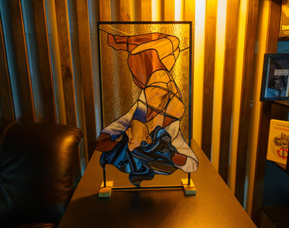 Stained glass abstract female figure panel displayed on table stand in warm ambient light, amber and navy glass tones, feminist fine art home decor