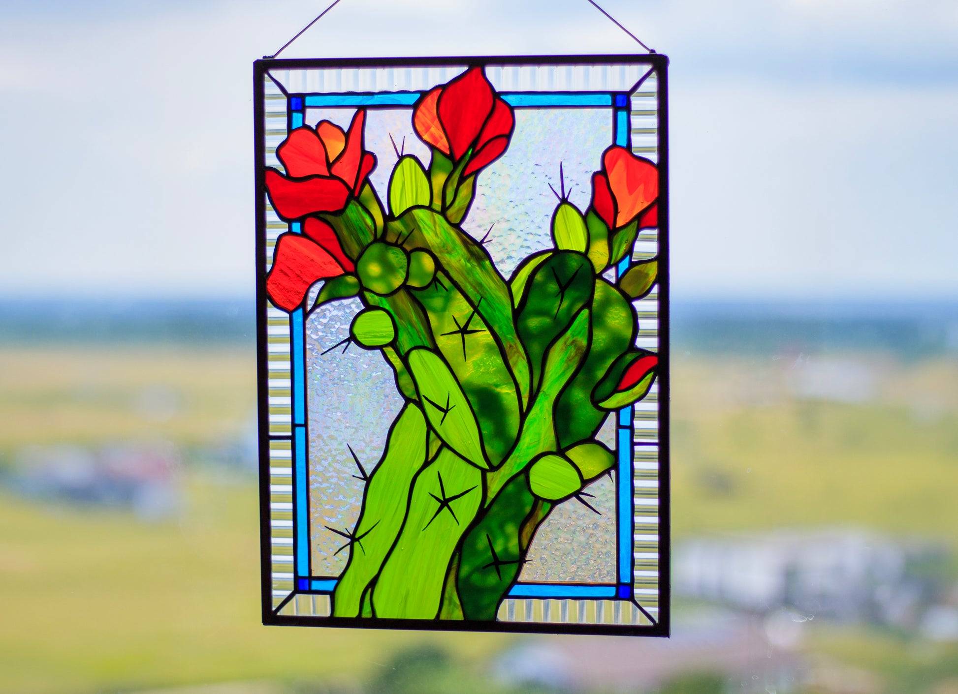 Blooming cactus stained glass panel hanging outdoors — green and red glass, blue border, copper foil Tiffany method, Mojoartglass