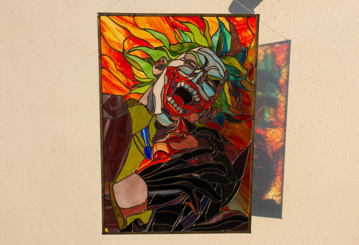 Creepy clown stained glass panel hanging on white wall in sunlight, large handmade copper foil horror comic art