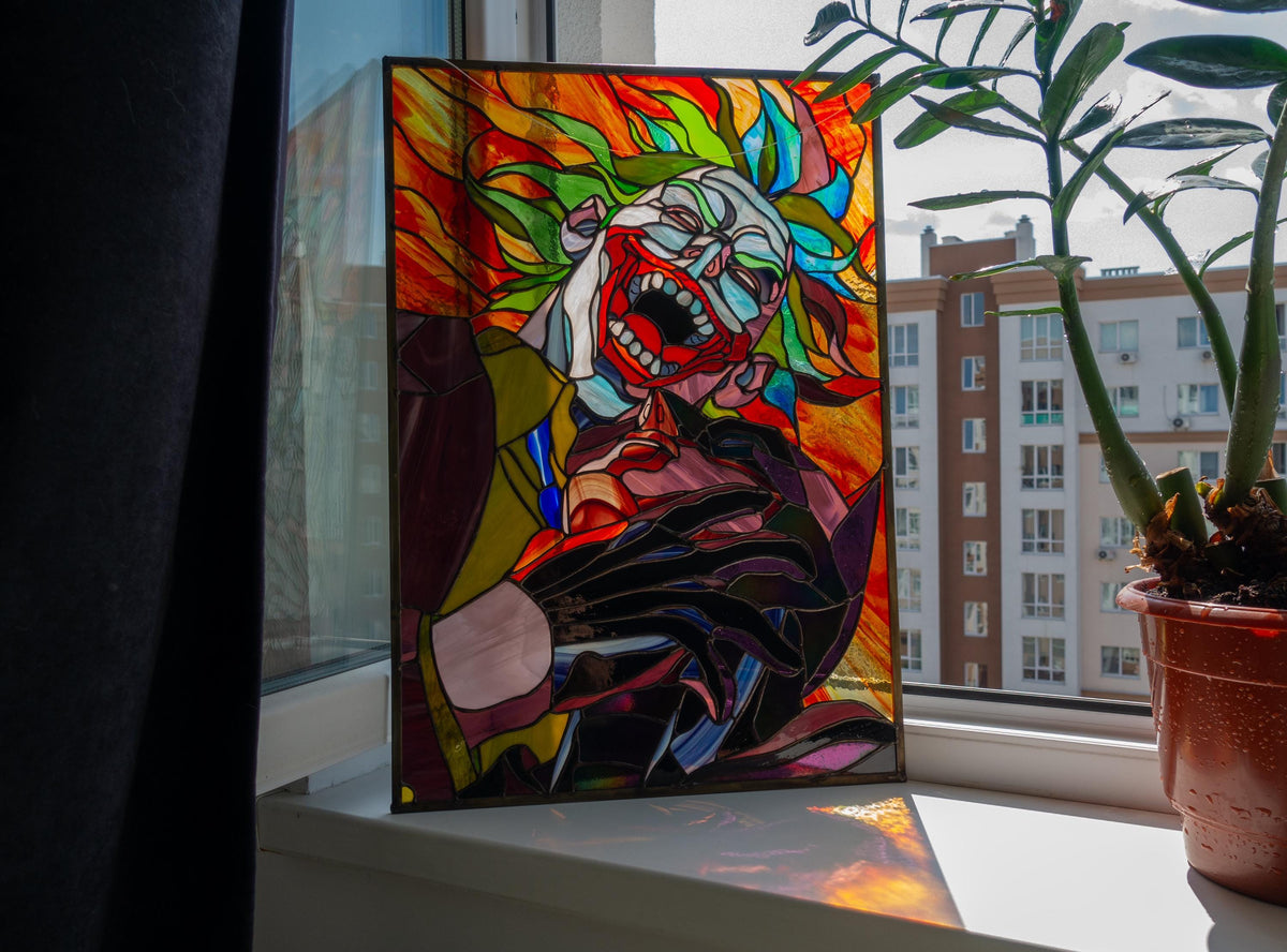 Creepy clown stained glass panel leaning on windowsill indoors, vibrant red orange green handmade Tiffany copper foil art