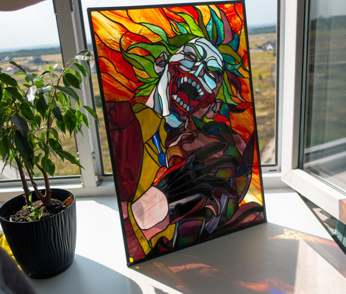 Creepy clown stained glass panel hanging in window with city view, backlit vibrant colors handmade Tiffany glass villain art