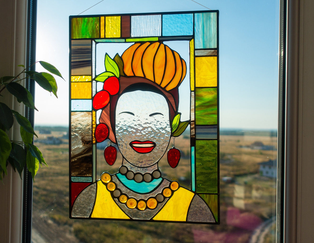 Stained glass panel of a woman with a colorful headdress and jewelry, placed on a window with a view of a field.