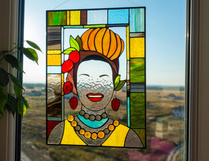 Stained glass panel of a woman with a colorful headdress and jewelry, placed on a window with a view of a field.