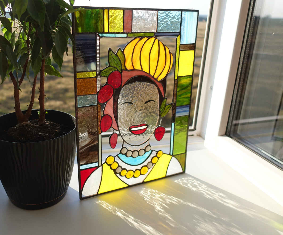 Stained glass panel of a woman with a yellow headscarf and red flowers, placed on a surface with a plant in the background.