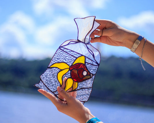 Goldfish in a bag stained glass suncatcher hanging in window — nostalgic carnival fish, warm orange glow, made in Ukraine