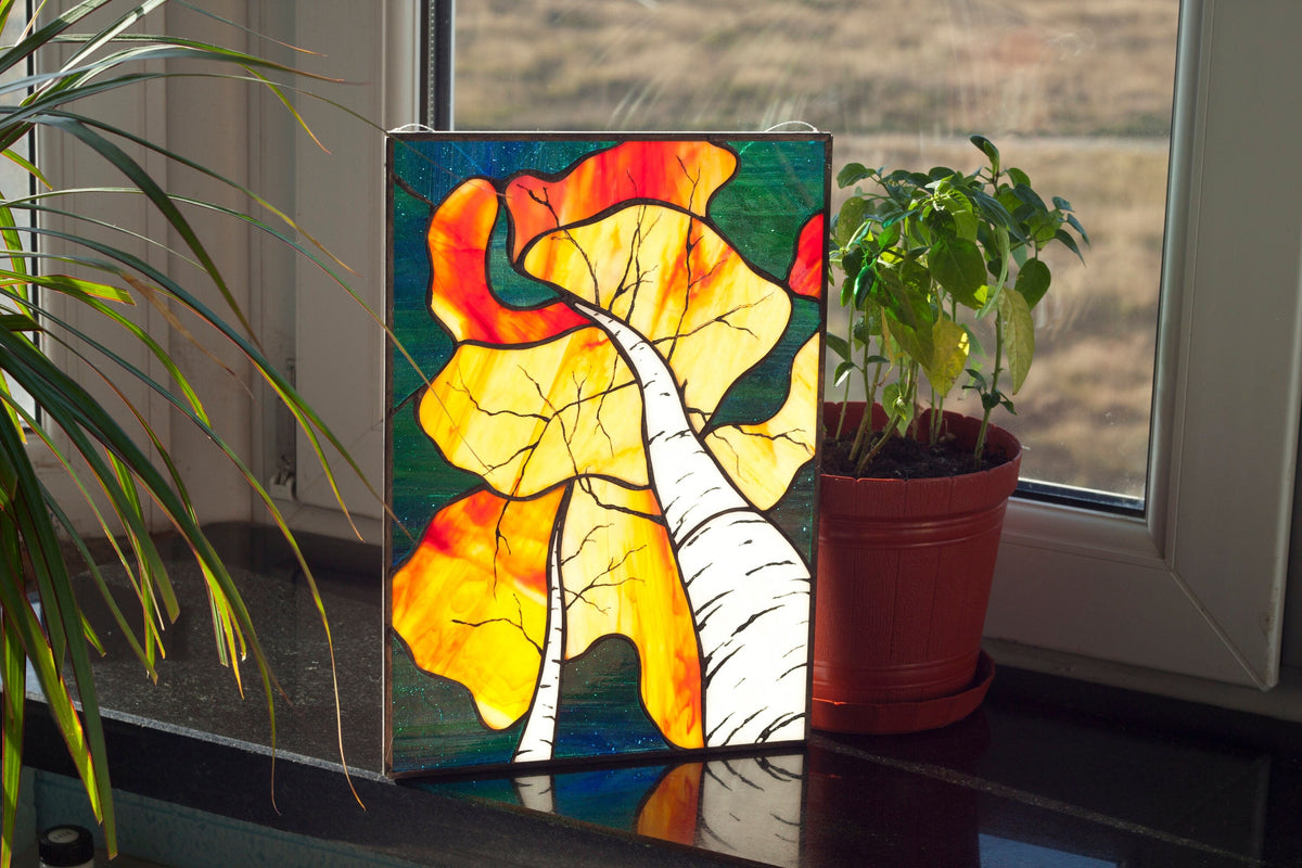 Handmade stained glass autumn birch forest panel standing on windowsill with plants, golden and orange foliage with white birch trunks, copper foil technique