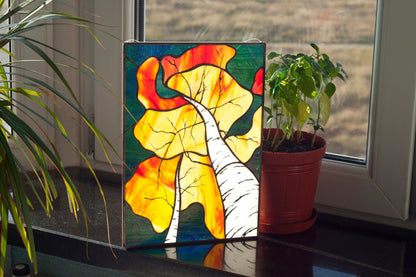 Handmade stained glass autumn birch forest panel standing on windowsill with plants, golden and orange foliage with white birch trunks, copper foil technique