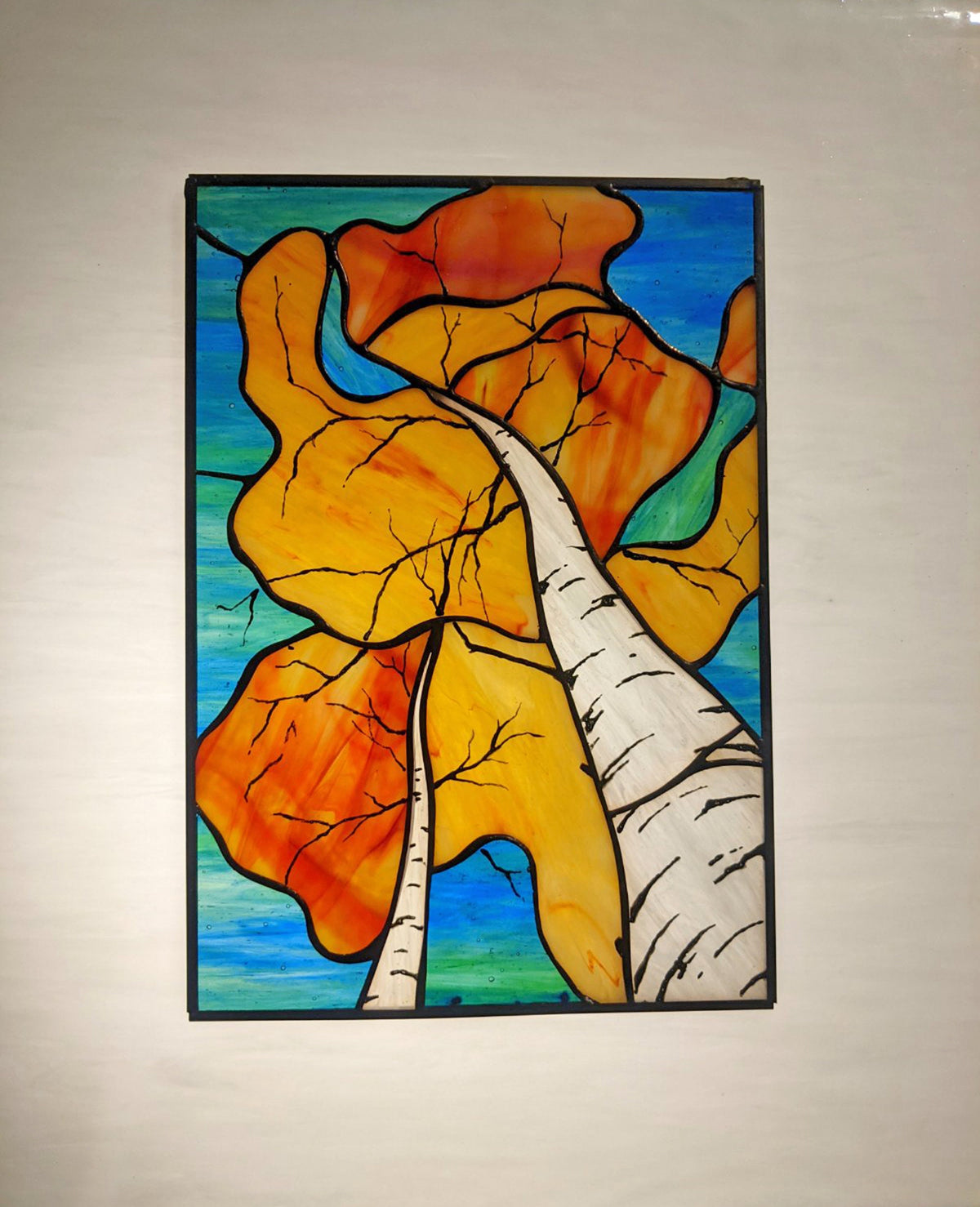Stained glass artwork of autumn leaves and a birch tree on a beige wall.