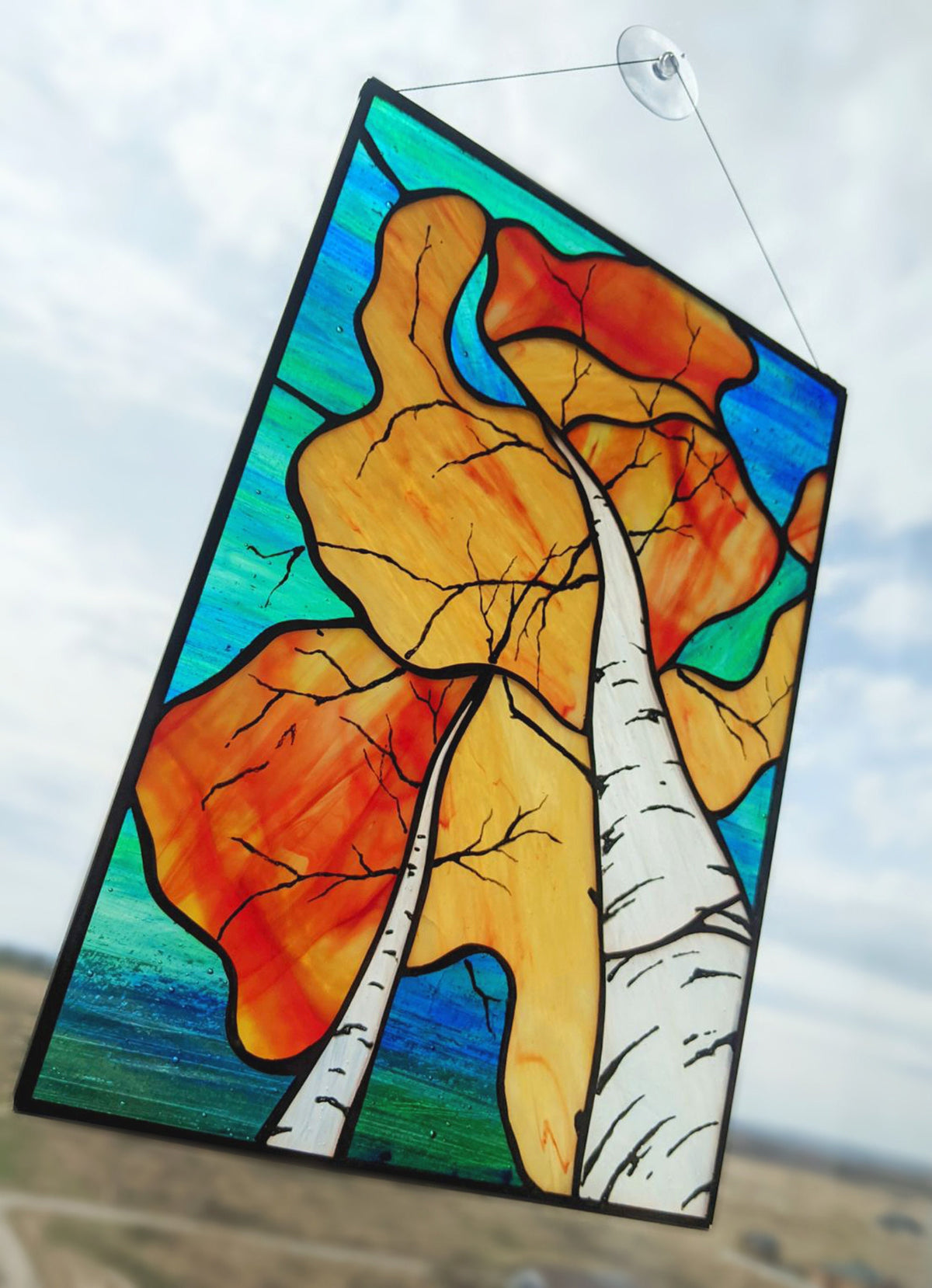 Stained glass panel with autumn leaves and a tree against a blue sky.