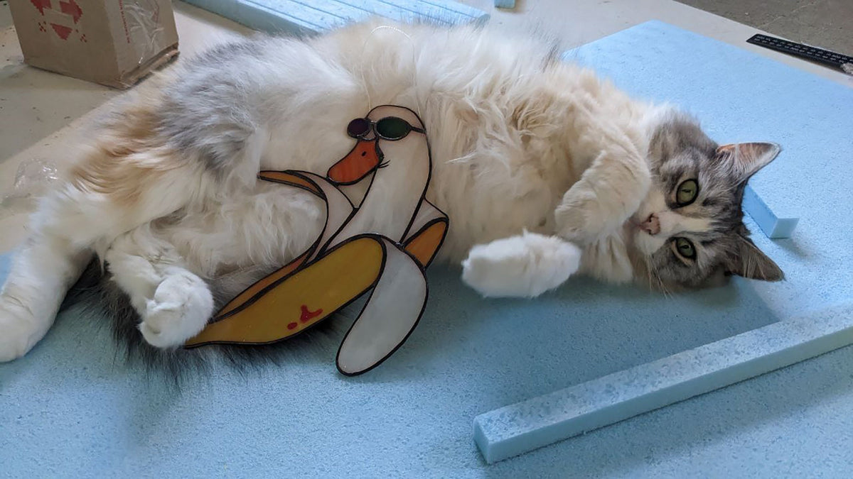 Fluffy cat lying next to handmade banana duck stained glass suncatcher on studio floor, funny lifestyle photo