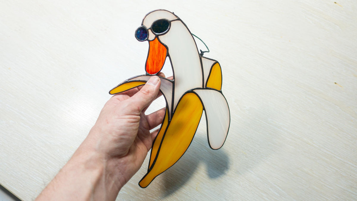 Handmade stained glass banana duck suncatcher with blue sunglasses held in hand against white background, showing full size and detail