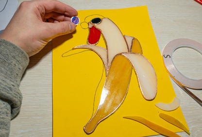 Work in progress — banana duck stained glass suncatcher being assembled on pattern, copper foil stage, blue glass sunglass piece held in hand