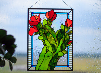 Handmade stained glass cactus panel in window — red blooms, green glass, blue frame border, plant lover wall art