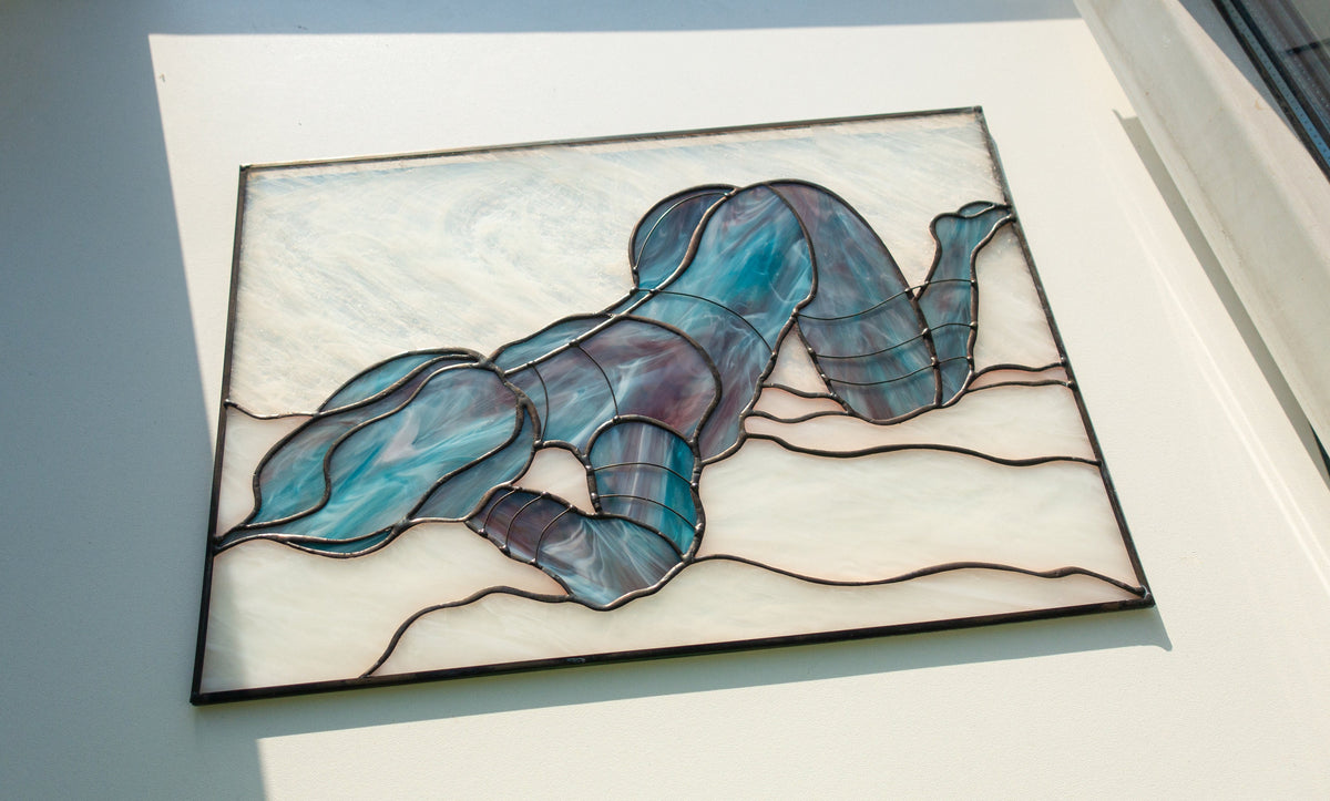Handmade stained glass reclining female figure panel hanging on window at sunset, iridescent blue and purple glass, warm orange glow through, copper foil Tiffany method