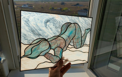 Hand holding large stained glass female figure panel up to window, minimalist line art nude in iridescent blue glass, landscape visible through glass background