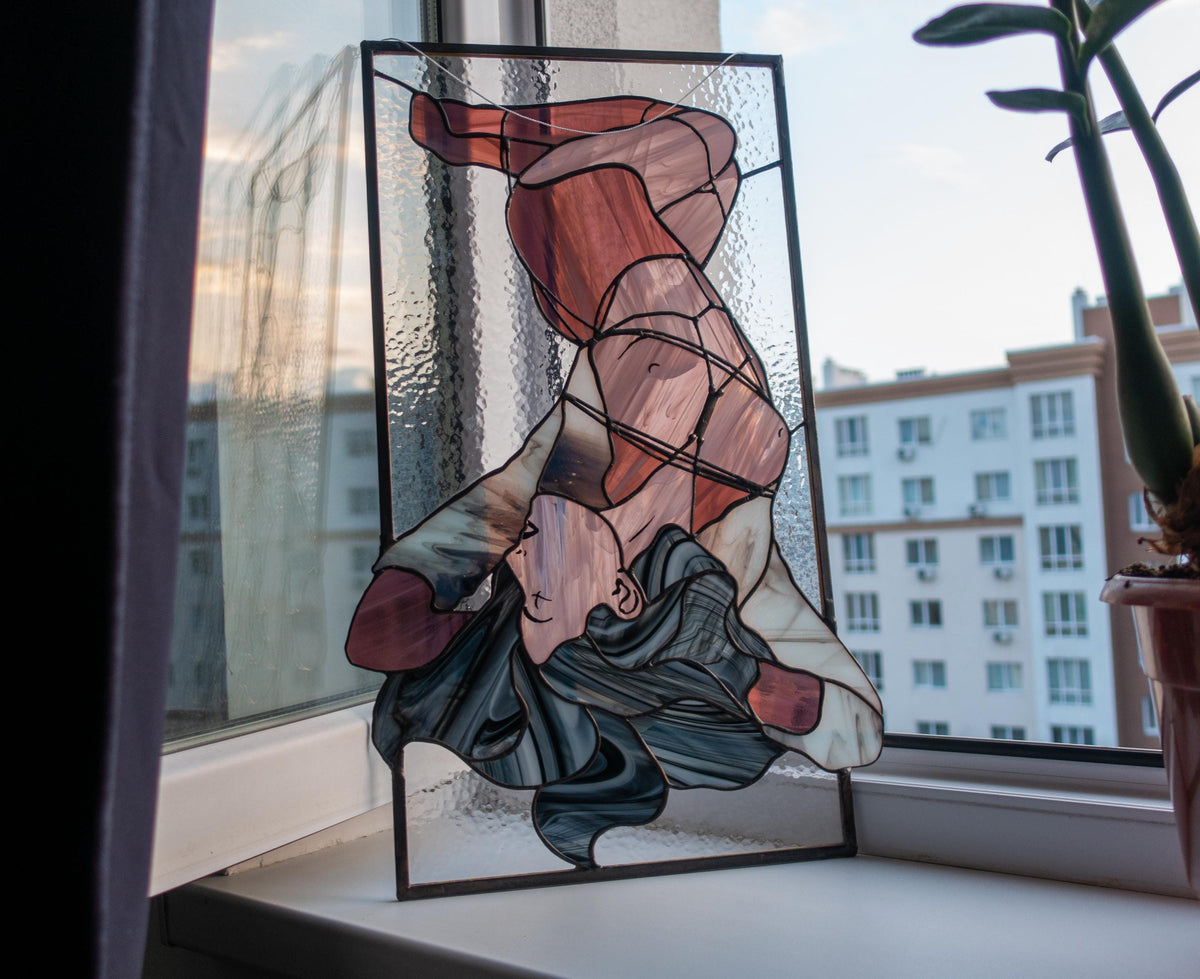 Abstract female figure stained glass panel leaning on windowsill at dusk, reclining nude in mauve pink and slate glass, plant visible beside, city view