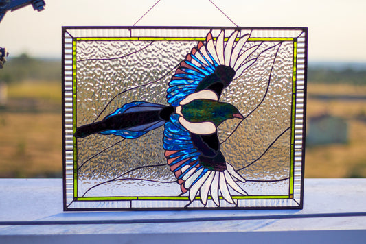 Handmade stained glass magpie panel held up against blue sky, iridescent blue and teal wings, red border frame, copper foil Tiffany method