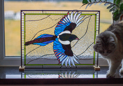 Stained glass flying magpie panel on window ledge with curious cat nearby, handmade bird art with lime border, home decor lifestyle photo