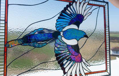 Iridescent magpie stained glass window panel displayed on windowsill, blue teal wings with red accent border, handmade bird art