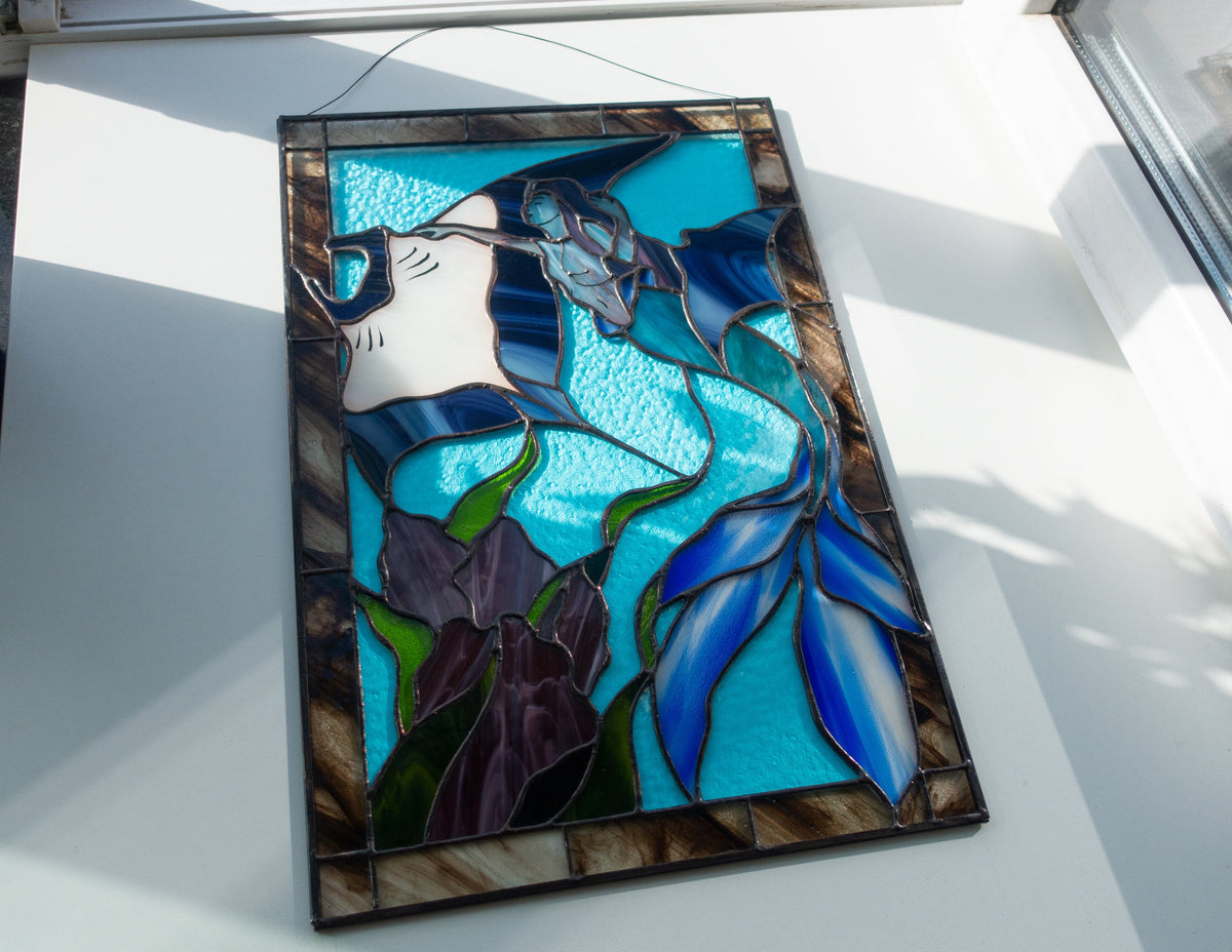 Handmade stained glass mermaid panel on white surface in sunlight, bright turquoise ocean glass with blue mermaid tail, white stingray and purple seaweed detail