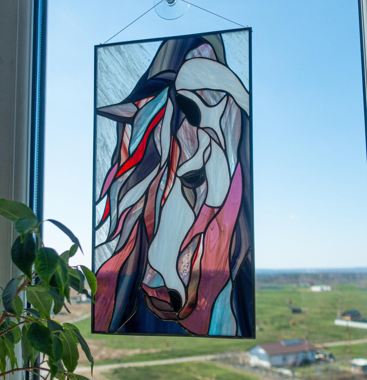 Handmade stained glass mustang horse panel hanging in a window with suction cup, blue sky background, abstract horse portrait in pink and blue glass