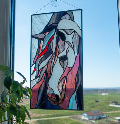 Handmade stained glass mustang horse panel hanging in a window with suction cup, blue sky background, abstract horse portrait in pink and blue glass