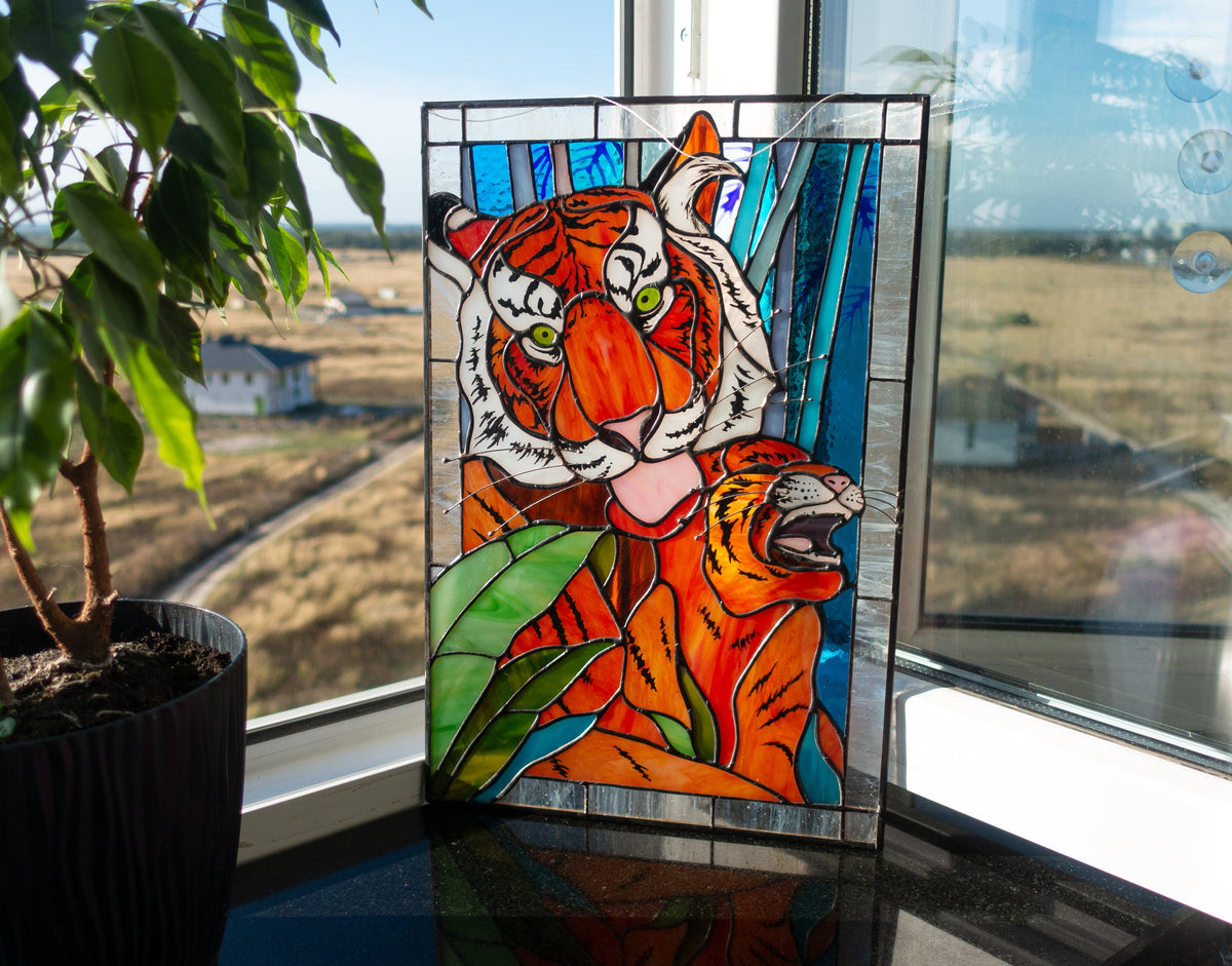 Stained glass panel of a tiger on a windowsill with a view outside.