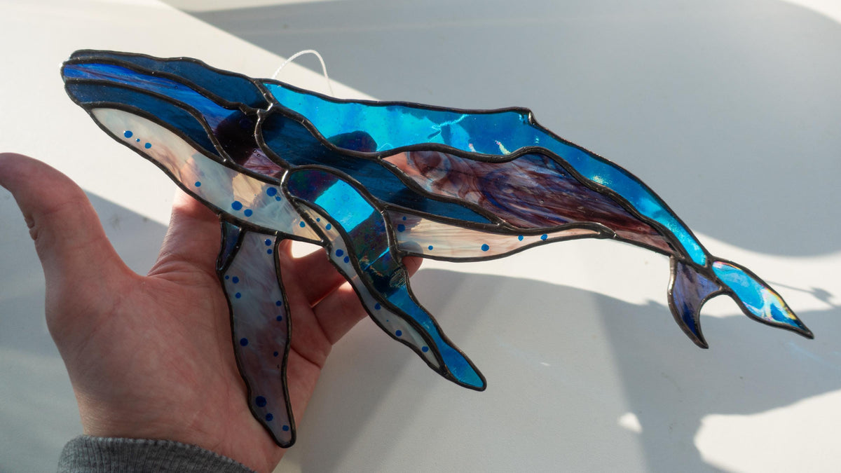 Handmade stained glass humpback whale suncatcher held in hand, blue and purple iridescent glass with white belly and copper foil detail, example of finished pattern result
